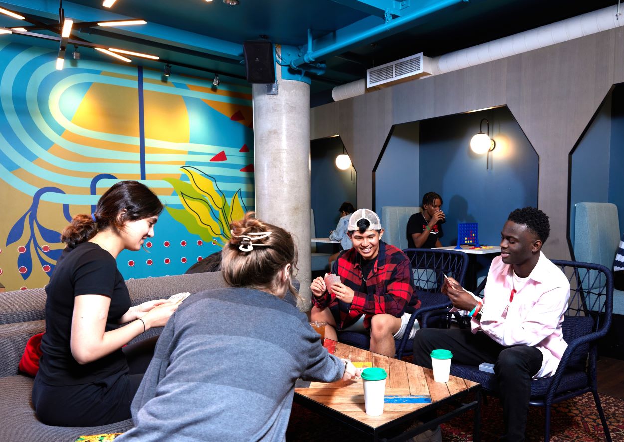 Four students play cards together at a table beside a colourful mural